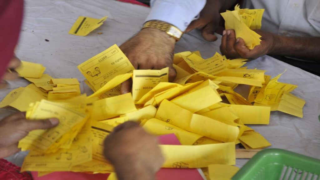 मतगणना के दौरान टेबल पर रखे बैलेट पेपर, जहां जांच अधिकारी मतपत्रों की जांच कर रहे हैं, चुनाव गड़बड़ी का प्रतीक दृश्य।